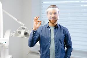 Portrait of handsome male dentist wearing in big protection medical shield on face preparing for dental surgery. Man smiling and looking at camera. photo