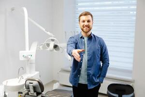 The happy adult man extends a hand of greetings, standing near stomatology chair in dentist office photo