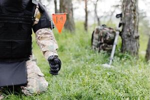 un hombre en un militar uniforme y un a prueba de balas chaleco trabajos en el bosque a desminar el territorio. un hombre advierte de peligro por haciendo un rojo marca foto