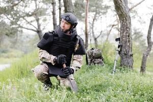 un hombre en un militar uniforme y un a prueba de balas chaleco trabajos en el bosque a desminar el territorio. un hombre excava explosivos en el bosque foto