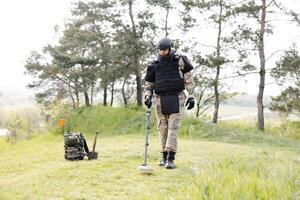 un hombre en un militar uniforme y a prueba de balas chaleco trabajos en el bosque con un metal detector. un dragaminas realiza trabajo en desminado el territorio foto