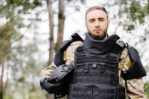 joven soldado en uniformes y táctico chaleco trabajos en el bosque y prepara para acción a un temporal bosque base. un hombre lo hace en el trabajo de desminado el territorio foto