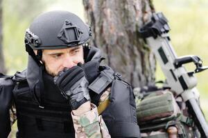 un hombre en un militar uniforme y un a prueba de balas chaleco se sienta en el bosque cerca un metal detector y un militar mochila. un hombre pausas en el trabajo de desminado el territorio foto