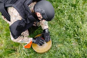 un hombre en un militar uniforme y un a prueba de balas chaleco trabajos en el bosque a desminar el territorio. un hombre advierte de peligro por haciendo un rojo marca foto