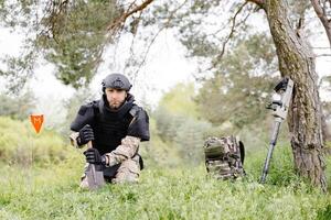 A man in a military uniform and a bulletproof vest works in the forest to demine the territory. A man warns of danger by making a red mark photo