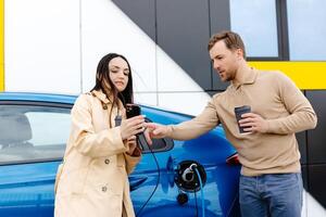 Young couple man and woman traveling by electric car having stop at charging station. Boyfriend plugging in cable to charge. Man talking with girlfriend, holding cup drinking hot coffee smiling photo