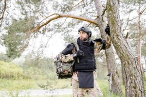 joven soldado en uniformes y táctico chaleco trabajos en el bosque y prepara para acción a un temporal bosque base. un hombre lo hace en el trabajo de desminado el territorio foto