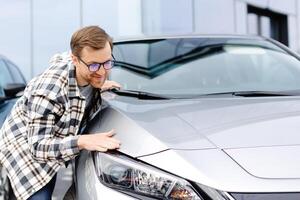 Young bearded man embracing his new car at the dealership smiling joyfully photo