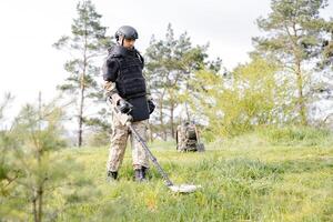 A man in a military uniform and bulletproof vest works in the forest with a metal detector. A minesweeper performs work on demining the territory photo