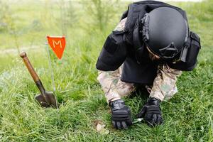 A man in a military uniform and a bulletproof vest works in the forest to demine the territory. A man warns of danger by making a red mark photo