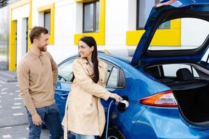 Young couple man and woman traveling by electric car having stop at charging station. Girlfriend holding charger looking at car and smiling photo