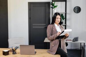 positivo mujer sentado a oficina mesa escritura notas en papel mientras trabajando en proyecto en moderno lugar de trabajo foto