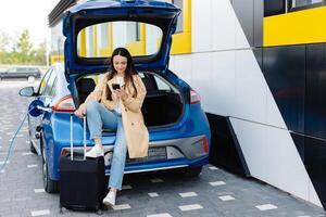 Young woman waiting charging automobile battery from small public station and using smartphone while charging automobile photo