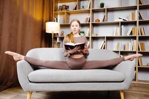 joven hermosa rubia mujer en marrón polainas y parte superior sentado en un enroscarse a hogar en el sofá. el niña es leyendo un libro sentado en el sofá en un raro posición foto