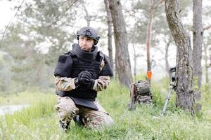 un hombre en un militar uniforme y un a prueba de balas chaleco trabajos en el bosque a desminar el territorio. un hombre pone en un protector casco antes de comenzando trabajo foto