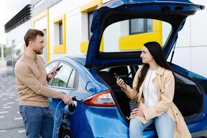 Young couple man and woman traveling by electric car having stop at charging station. Boyfriend plugging in cable to charge. Man talking with girlfriend, holding cup drinking hot coffee smiling photo