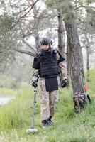 A man in a military uniform and bulletproof vest works in the forest with a metal detector. A minesweeper performs work on demining the territory photo