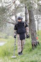 un hombre en un militar uniforme y a prueba de balas chaleco trabajos en el bosque con un metal detector. un dragaminas realiza trabajo en desminado el territorio foto