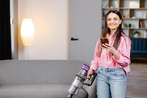 Happy cheerful young Caucasian beautiful woman vacuuming floor and texting, surfing internet on smartphone at home in living room. Pretty female using vacuum cleaner, domestic concept photo