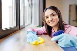 mujer en protector guantes es sonriente y limpiando polvo utilizando un rociar y un plumero mientras limpieza su casa, de cerca foto