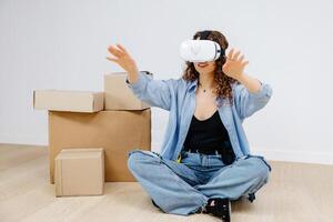 Young woman in VR glasses using modern technology creating room design, moving hands in air, sitting in new apartment during renovation. Virtual reality and repair concept. photo