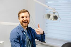 linda joven hombre sentado en un silla en un hermosa moderno dental oficina, sonriente y demostración dedos gesto genial trabajo foto