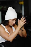 retrato confidente hermosa joven morena mujer es vistiendo blanco traje y gorra sentado en el gimnasio foto