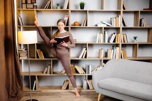 joven mujer en ropa de deporte extensión piernas, haciendo divisiones en pie en biblioteca y leyendo libro. hembra en diario rutina de ejercicio hacer yoga formación a hogar foto