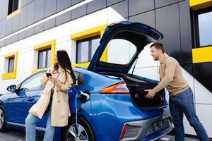 Young couple man and woman traveling by electric car having stop at charging station. The man taking a suitcase from the trunk. Man talking with girlfriend and she holding mobile phone in hand photo