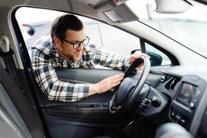 Young man looking through window inside new car, checking it before purchase at modern dealership store. Cheerful guy buying or renting automobile at showroom photo