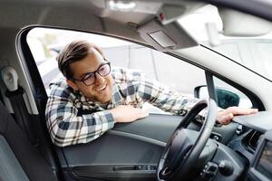 Young man looking through window inside new car, checking it before purchase at modern dealership store. Cheerful guy buying or renting automobile at showroom photo