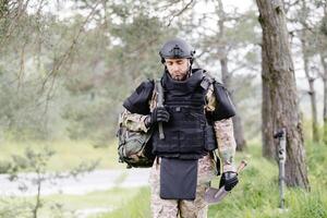 joven soldado en uniformes y táctico chaleco trabajos en el bosque y prepara para acción a un temporal bosque base. un hombre lo hace en el trabajo de desminado el territorio foto