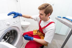 el joven hermoso reparador en trabajador traje con el profesional herramientas caja es fijación el Lavado máquina en el baño. foto