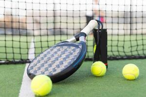 azul padel raqueta y amarillo pelotas metido en Corte cerca red en soleado día foto