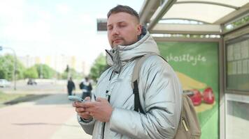 Young attractive bearded Caucasian man standing on the bus station, waiting public transport, looking around, watching at phone screen and reading news information. video