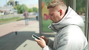 ung attraktiv skäggig caucasian man stående på de buss station, väntar offentlig transport, ser runt om, tittar på på telefon skärm och läsning Nyheter information. video