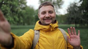 freddo barbuto caucasico uomo in piedi nel natura parco utilizzando mobile Telefono. ritratto di sorridente giovane maschio turista fabbricazione un' autoscatto o chiamata a verde giungla foresta sfondo. video