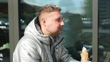 Portrait of handsome young Caucasian man eating tasty hot dog and drinking coffee sitting in outdoors cafe. Delivery service worker with tasty food video