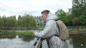 joven hermoso caucásico chico camina a primavera parque, mecanografía mensaje, utilizando móvil teléfono aplicación fuera de cerca un ciudad lago video