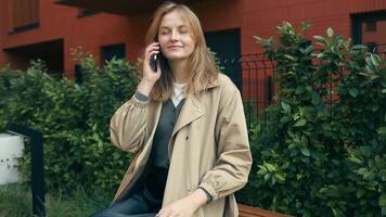 ritratto di elegante sorridente attività commerciale donna nel alla moda Abiti chiamata su mobile Telefono, si siede su panchina nel città strada video