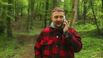 joven hipster hombre en casual ropa llamadas en el teléfono soportes en el bosque, comunica en un teléfono inteligente caucásico viajero con un mochila turista haciendo un llamada soportes a el primavera bosque video