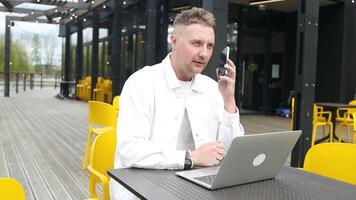 Happy successful European businessman makes a phone call on smartphone, talking and smiling outdoors sits at city cafe on city street outside. video