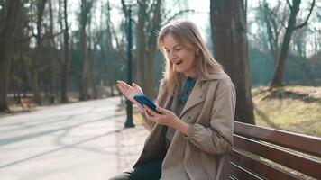 Excited happy blonde woman receiving good news on smartphone outdoors show winner gesture, sitting on the bench at spring park video