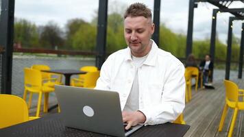 Side view of Caucasian man who sits in city outdoors cafe in daytime and using the laptop for the remote work. Freelancer businessman typing text on the keyboard in coffeeshop video
