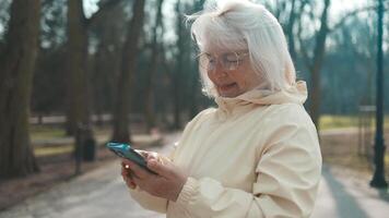 Joyful elderly senior active woman with a phone in her hands enjoys outdoor recreation. Lady looking at smart phone, while sitting on the bench in the park. Using modern technology video
