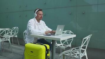 Style travel man uses laptop computer, listen to music or podcast in headphone, waiting for flight. Traveling digital remote work online in boarding lounge of airline hub video