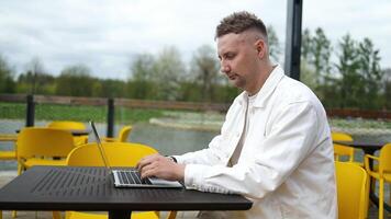 Side view of Caucasian man who sits in city outdoors cafe in daytime and using the laptop for the remote work. Freelancer businessman typing text on the keyboard in coffeeshop video