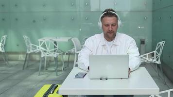 Friendly Caucasian 30s man sits in airport terminal using laptop talking via call. Male waiting for flight, browse internet, social media, online shopping. video