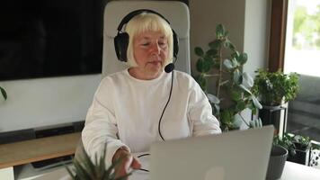 senior 60s attraktiv influencer kvinna bär hörlurar och en leende använder sig av bärbar dator, inlärning språk, tittar på uppkopplad webbseminarium, lyssnande audio kurs, e-learning utbildning video