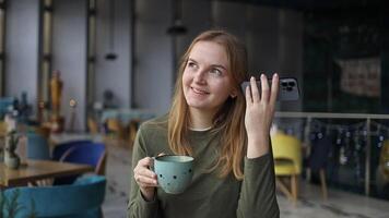 Pretty female student listens autodidact sms and smartphone browses internet rests in cozy cafe has break at daytime drinks coffee. video